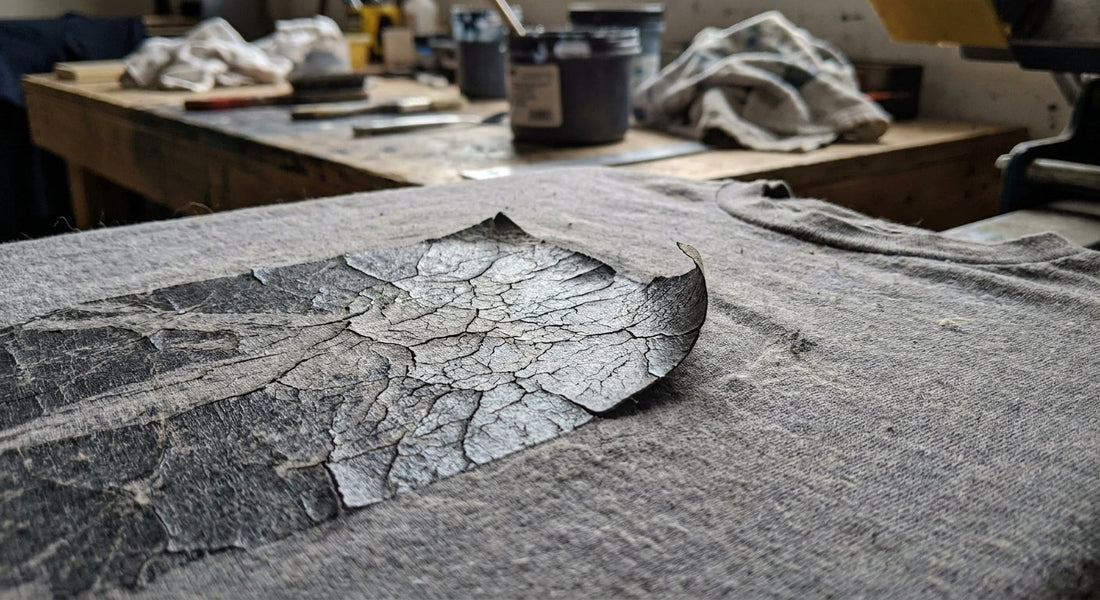 Macro close-up of a printed graphic on a worn grey t-shirt showing hairline cracks and one lifting edge