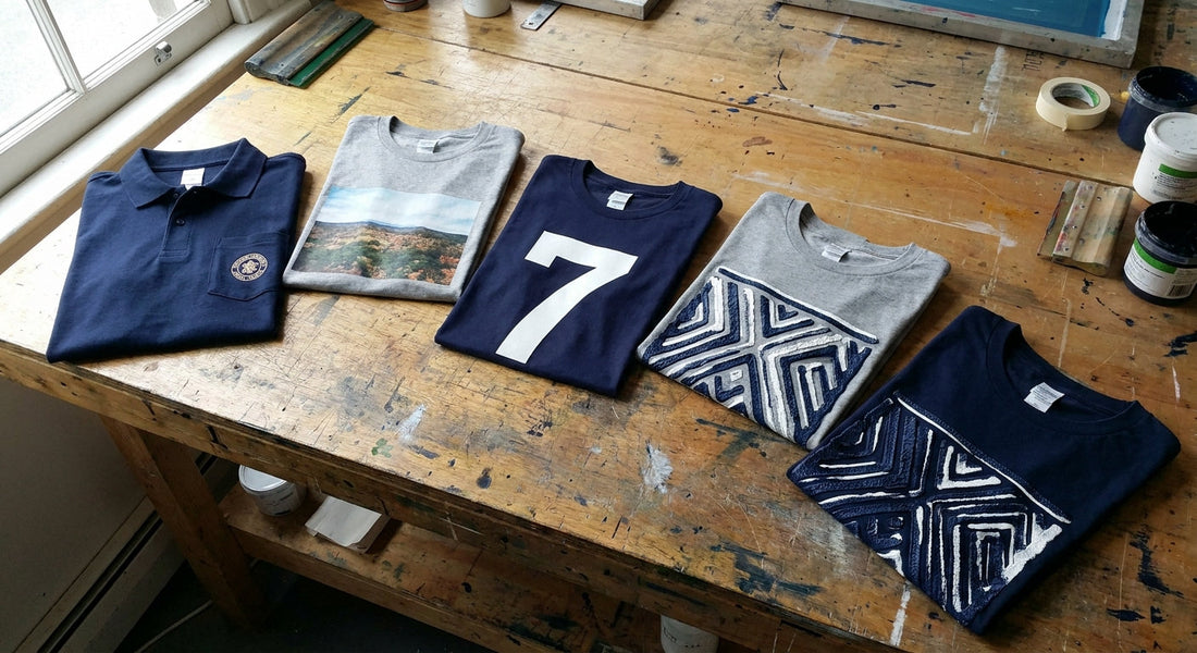 Four decorated t-shirts and a polo shirt laid out on a wooden table, each showing a different apparel decoration method